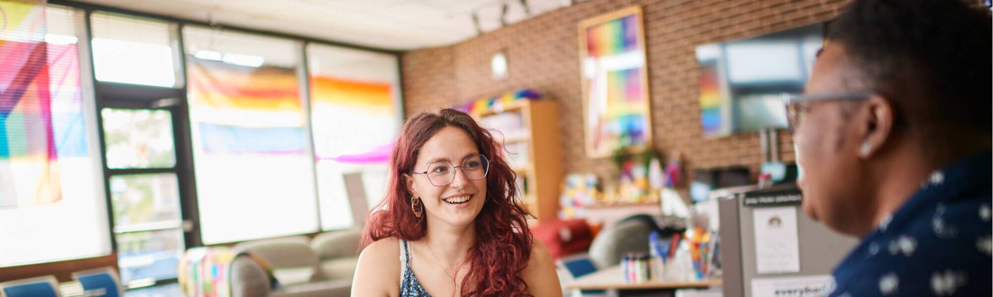 Two individuals engage in conversation at a table in a brightly lit, colorful room with rainbow flags and creative decor.